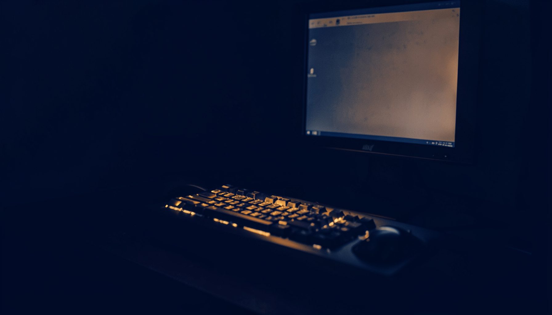 A deep navy and amber duotone photograph of a dimly lit desk with a keyboard and monitor, warm amber highlights on the keys and screen glow against dark surroundings
