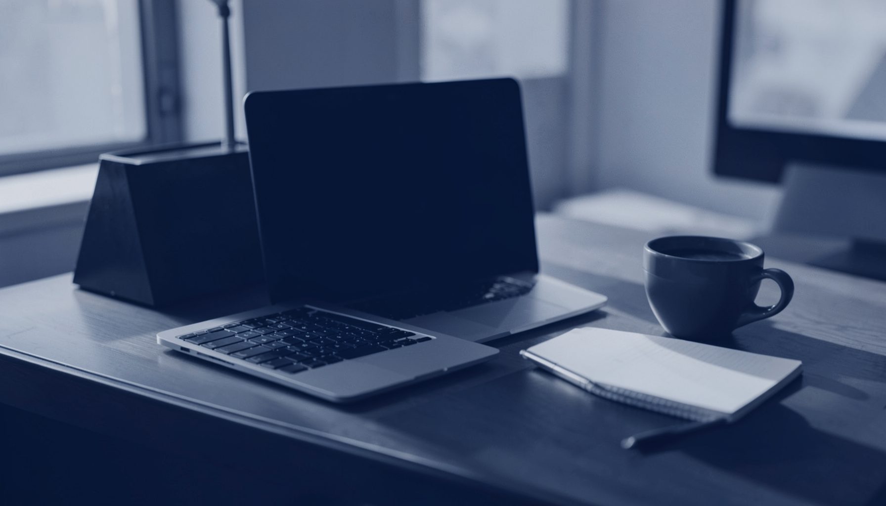 A deep indigo duotone photograph of a modern office desk with a laptop, notebook, and coffee cup, bathed in soft morning light.